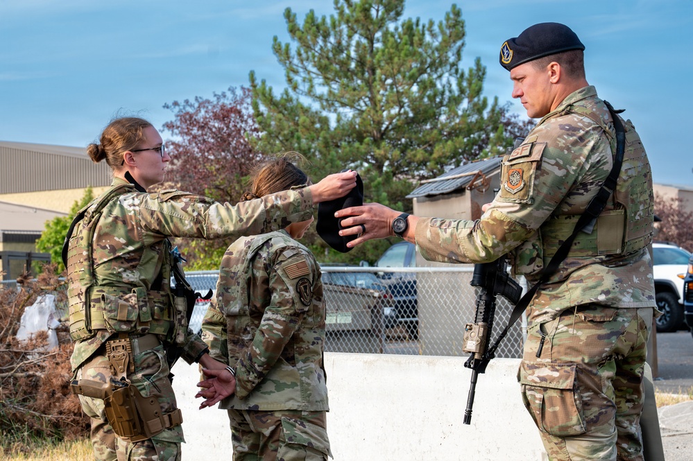 Three military personnel in OCP (Operational Camouflage Pattern) uniforms are engaged in what appears to be a training or security forces exercise. A female airman on the left is handing or exchanging a dark item (likely a hood or blindfold) with a male airman on the right who is wearing a black beret and carrying a rifle, while a third person stands between them with hands behind their back. The setting appears to be an outdoor military installation or base.