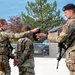 Three military personnel in OCP (Operational Camouflage Pattern) uniforms are engaged in what appears to be a training or security forces exercise. A female airman on the left is handing or exchanging a dark item (likely a hood or blindfold) with a male airman on the right who is wearing a black beret and carrying a rifle, while a third person stands between them with hands behind their back. The setting appears to be an outdoor military installation or base.
