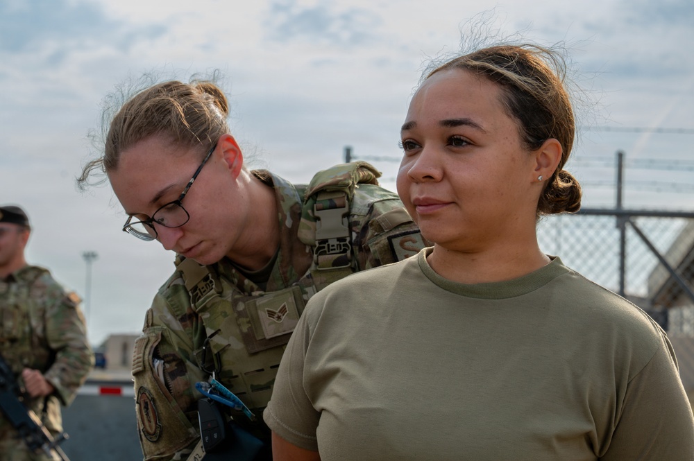 Two female military personnel are shown in an outdoor setting, likely on a military installation. One individual in full OCP (Operational Camouflage Pattern) uniform with tactical gear appears to be conducting a check or inspection on the second individual wearing an olive drab t-shirt. A chain-link fence with barbed wire and additional uniformed personnel are visible in the background.