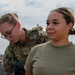 Two female military personnel are shown in an outdoor setting, likely on a military installation. One individual in full OCP (Operational Camouflage Pattern) uniform with tactical gear appears to be conducting a check or inspection on the second individual wearing an olive drab t-shirt. A chain-link fence with barbed wire and additional uniformed personnel are visible in the background.