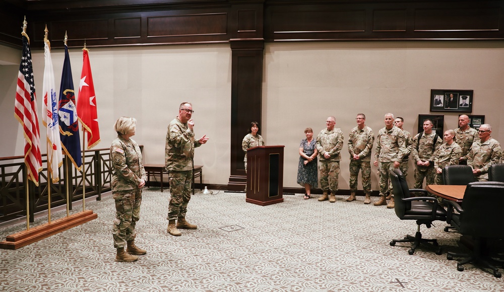 A military ceremony is taking place in a formal indoor setting, likely a conference or briefing room. A male soldier appears to be speaking to a female soldier standing before him, while a group of military personnel and at least one civilian observe from the background. Flags including the U.S. flag and what appears to be a general officer's flag are displayed prominently.