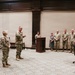A military ceremony is taking place in a formal indoor setting, likely a conference or briefing room. A male soldier appears to be speaking to a female soldier standing before him, while a group of military personnel and at least one civilian observe from the background. Flags including the U.S. flag and what appears to be a general officer's flag are displayed prominently.