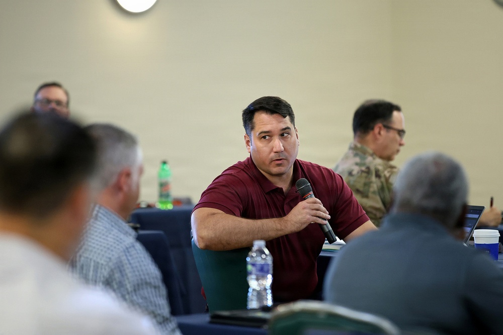 A man in a maroon polo shirt is speaking into a handheld microphone during what appears to be a formal meeting or conference. Several other attendees are visible, including one person in military camouflage uniform in the background. The setting suggests a structured briefing or panel discussion environment.
