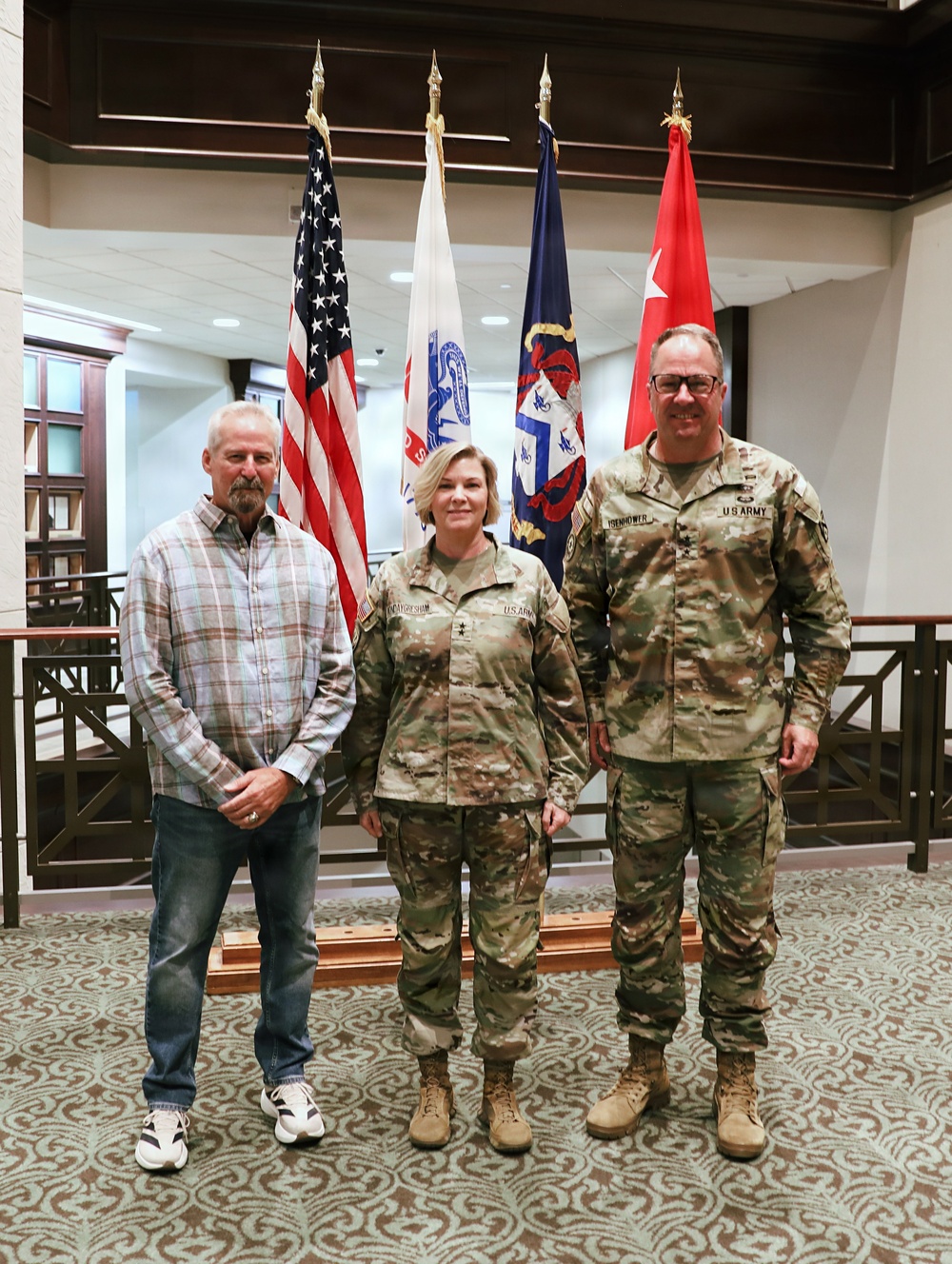 Three individuals pose for a formal photograph in what appears to be a U.S. Army facility. Two of the individuals are wearing U.S. Army OCP (Operational Camouflage Pattern) uniforms, while the third is in civilian clothing. Multiple military flags are displayed prominently in the background.
