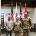 Three individuals pose for a formal photograph in what appears to be a U.S. Army facility. Two of the individuals are wearing U.S. Army OCP (Operational Camouflage Pattern) uniforms, while the third is in civilian clothing. Multiple military flags are displayed prominently in the background.