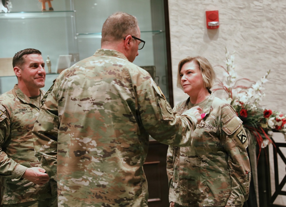 A military award ceremony is taking place indoors, where a senior officer is pinning a medal onto a female soldier in U.S. Army OCP (Operational Camouflage Pattern) uniform. A third soldier, identified by a name tape reading 'CURRY,' observes the ceremony from the left. The setting appears to be an official military venue with decorative floral arrangements visible in the background.