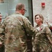 A military award ceremony is taking place indoors, where a senior officer is pinning a medal onto a female soldier in U.S. Army OCP (Operational Camouflage Pattern) uniform. A third soldier, identified by a name tape reading 'CURRY,' observes the ceremony from the left. The setting appears to be an official military venue with decorative floral arrangements visible in the background.