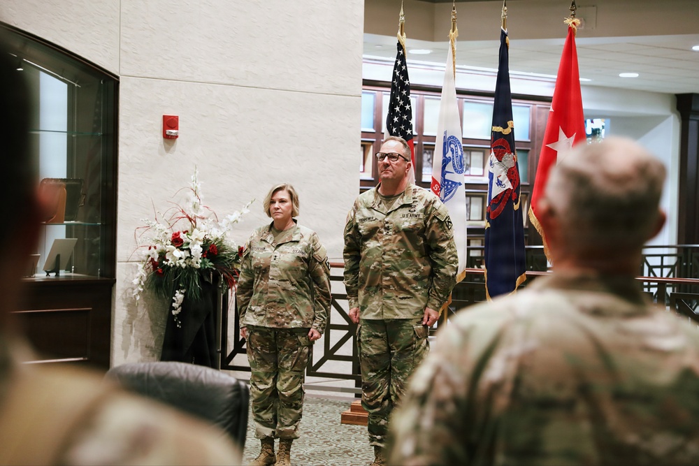 Two U.S. Army soldiers in MultiCam/OCP camouflage uniforms stand at attention during what appears to be a formal military ceremony held indoors. Multiple flags are displayed in the background, including the U.S. flag and what appears to be a general officer's flag (red with stars). An audience of uniformed personnel is visible in the foreground.