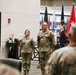 Two U.S. Army soldiers in MultiCam/OCP camouflage uniforms stand at attention during what appears to be a formal military ceremony held indoors. Multiple flags are displayed in the background, including the U.S. flag and what appears to be a general officer's flag (red with stars). An audience of uniformed personnel is visible in the foreground.