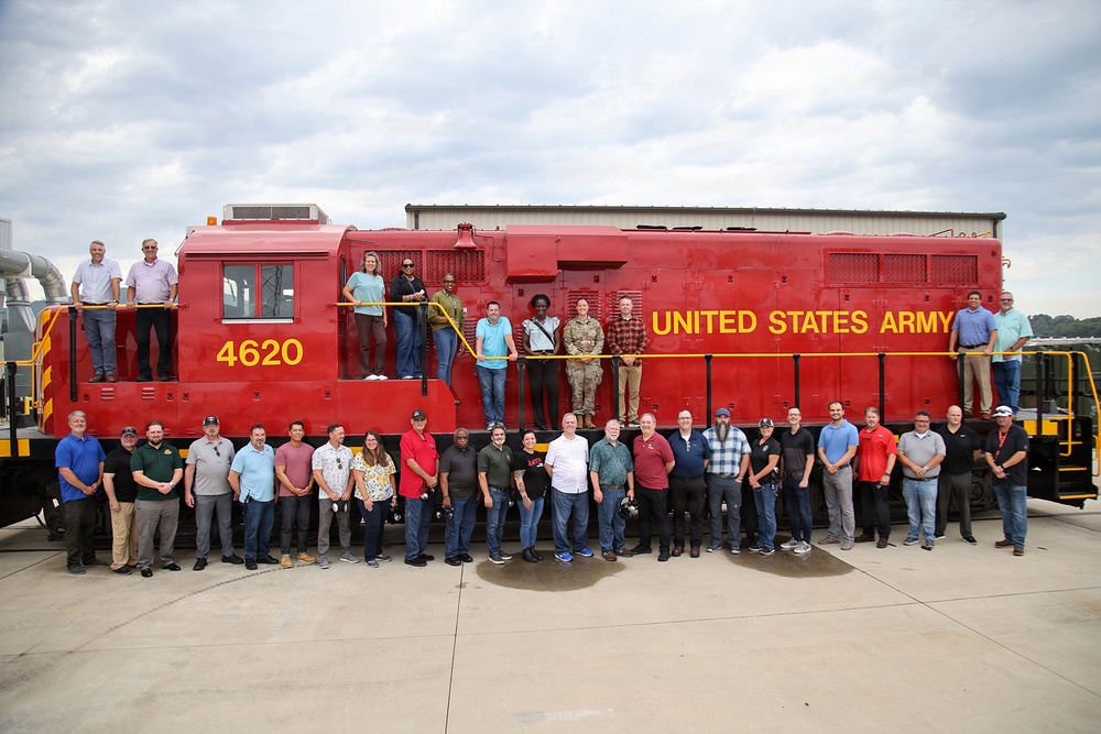 A large group of approximately 30 people poses in front of a bright red United States Army locomotive numbered 4620. Some individuals are standing on the locomotive's running boards and walkways while others stand on the ground in front of it. The photo appears to be an official group photograph taken at a military or industrial facility.