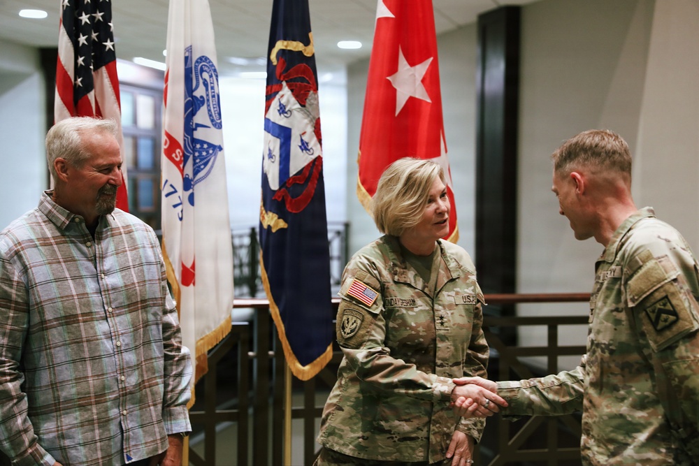 Two U.S. Army soldiers in OCP (Operational Camouflage Pattern) uniforms are shaking hands in what appears to be a formal military building, while a civilian man in a plaid shirt observes. Several military flags are displayed in the background, including the U.S. national flag and a general officer flag (red with white stars). The setting suggests an official military ceremony or meeting.