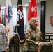 Two U.S. Army soldiers in OCP (Operational Camouflage Pattern) uniforms are shaking hands in what appears to be a formal military building, while a civilian man in a plaid shirt observes. Several military flags are displayed in the background, including the U.S. national flag and a general officer flag (red with white stars). The setting suggests an official military ceremony or meeting.