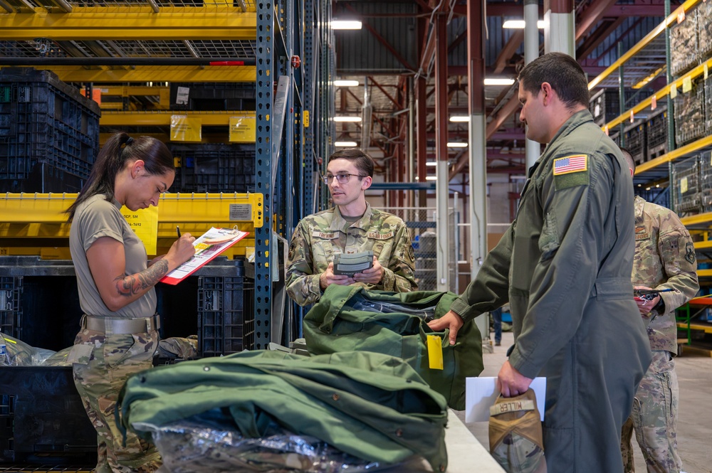 Military personnel are conducting what appears to be an equipment issue or inventory process inside a large warehouse or supply depot. Three primary individuals are visible: one taking notes on a clipboard, one holding a scanning device while managing gear, and one in a flight suit receiving or reviewing items. Green military gear and clothing items are laid out on a table in the foreground.