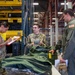 Military personnel are conducting what appears to be an equipment issue or inventory process inside a large warehouse or supply depot. Three primary individuals are visible: one taking notes on a clipboard, one holding a scanning device while managing gear, and one in a flight suit receiving or reviewing items. Green military gear and clothing items are laid out on a table in the foreground.