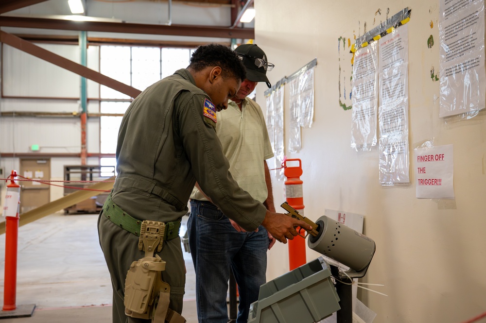A military or law enforcement personnel in an olive green flight suit is demonstrating proper firearm handling to a civilian at what appears to be a weapons clearing station or range facility. The uniformed individual is inserting a pistol into a clearing barrel, a standard safety procedure used to safely unload and verify firearms. A safety sign reading 'FINGER OFF THE TRIGGER!!!' is prominently posted on the wall nearby.