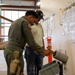 A military or law enforcement personnel in an olive green flight suit is demonstrating proper firearm handling to a civilian at what appears to be a weapons clearing station or range facility. The uniformed individual is inserting a pistol into a clearing barrel, a standard safety procedure used to safely unload and verify firearms. A safety sign reading 'FINGER OFF THE TRIGGER!!!' is prominently posted on the wall nearby.