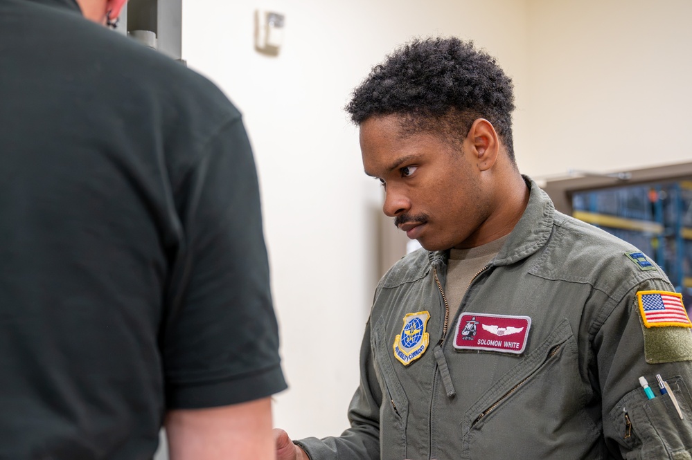 A U.S. Air Force service member named Solomon White, wearing an olive green flight suit, is engaged in a focused interaction with another individual whose back is to the camera. His flight suit displays an Air Mobility Command patch, a name tag with navigator/aircrew wings, and a U.S. flag patch on the sleeve. The setting appears to be an indoor military facility.