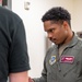 A U.S. Air Force service member named Solomon White, wearing an olive green flight suit, is engaged in a focused interaction with another individual whose back is to the camera. His flight suit displays an Air Mobility Command patch, a name tag with navigator/aircrew wings, and a U.S. flag patch on the sleeve. The setting appears to be an indoor military facility.