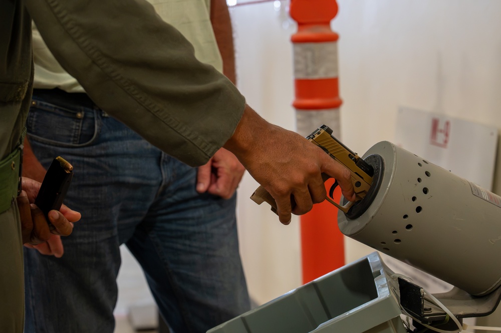 A person in a green jacket and jeans is inserting a gold/tan-colored pistol into what appears to be a gray cylindrical firearm clearing/safety device, commonly used at security checkpoints or shooting ranges. A second individual is visible in the background holding what appears to be a magazine. The setting appears to be an indoor facility with an orange safety cone visible in the background.