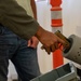 A person in a green jacket and jeans is inserting a gold/tan-colored pistol into what appears to be a gray cylindrical firearm clearing/safety device, commonly used at security checkpoints or shooting ranges. A second individual is visible in the background holding what appears to be a magazine. The setting appears to be an indoor facility with an orange safety cone visible in the background.