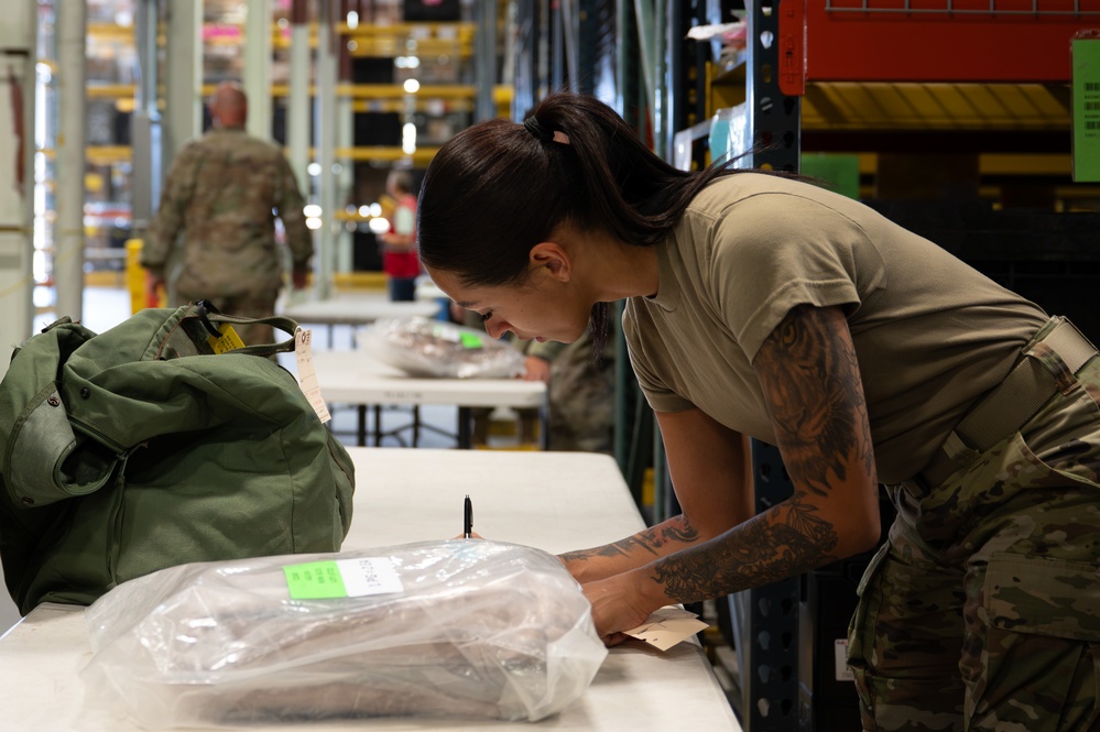 A female military service member in OCP (Operational Camouflage Pattern) uniform is leaning over a table in what appears to be a military supply warehouse or logistics facility. She is examining or processing a plastic-wrapped package with a green label, alongside a military duffel bag. Additional personnel and warehouse shelving are visible in the background.