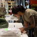 A female military service member in OCP (Operational Camouflage Pattern) uniform is leaning over a table in what appears to be a military supply warehouse or logistics facility. She is examining or processing a plastic-wrapped package with a green label, alongside a military duffel bag. Additional personnel and warehouse shelving are visible in the background.