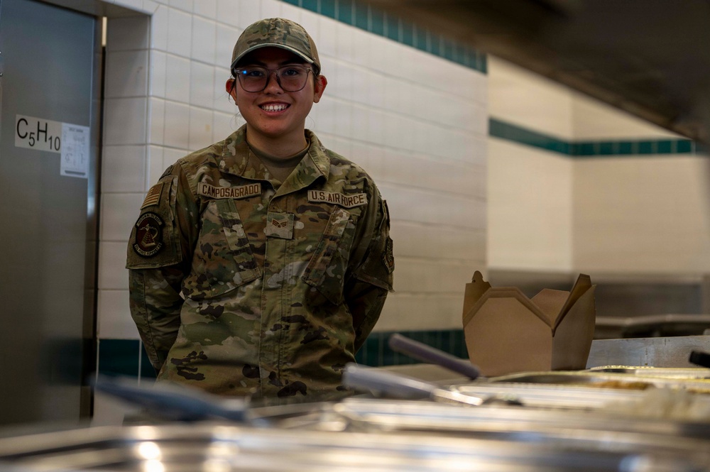 A U.S. Air Force Airman named Camposagrado is smiling while working behind a food service counter in what appears to be a military dining facility (DFAC). The airman is wearing an OCP (Operational Camouflage Pattern) uniform with visible rank insignia and U.S. Air Force tape. The setting suggests food service or mess hall duty.