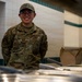 A U.S. Air Force Airman named Camposagrado is smiling while working behind a food service counter in what appears to be a military dining facility (DFAC). The airman is wearing an OCP (Operational Camouflage Pattern) uniform with visible rank insignia and U.S. Air Force tape. The setting suggests food service or mess hall duty.