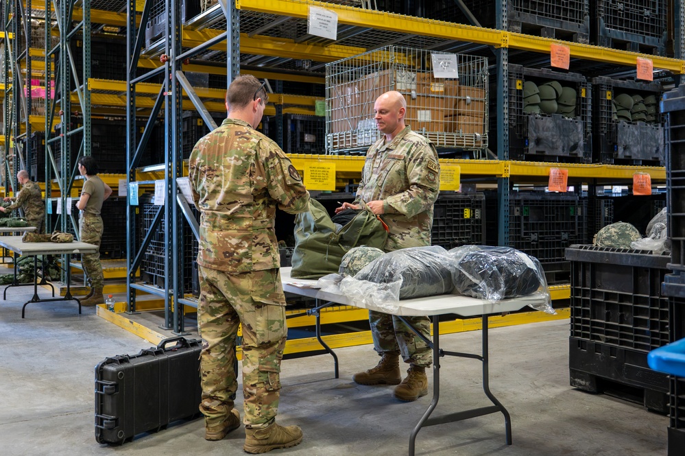 Two military personnel in OCP (Operational Camouflage Pattern) uniforms are interacting across a folding table in a large military supply warehouse. The facility contains tall yellow-framed shelving units stocked with military equipment including helmets and gear. A third service member is visible in the background working at another table.