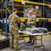 Two military personnel in OCP (Operational Camouflage Pattern) uniforms are interacting across a folding table in a large military supply warehouse. The facility contains tall yellow-framed shelving units stocked with military equipment including helmets and gear. A third service member is visible in the background working at another table.