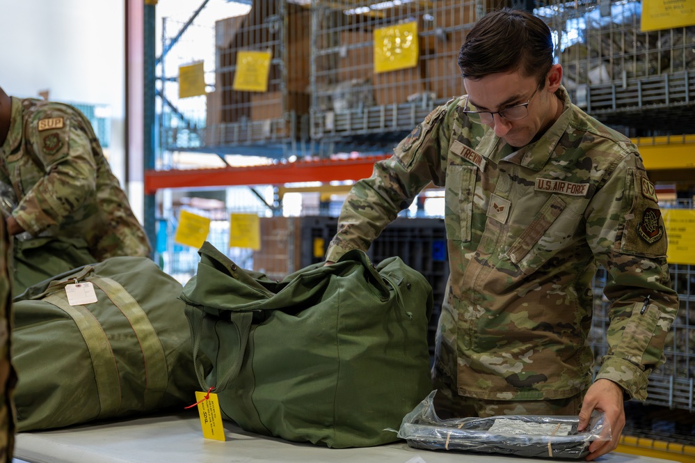 A U.S. Air Force airman named Trevor is working in what appears to be a supply or logistics warehouse, handling military duffel bags and packaged equipment on a work table. The airman is wearing OCP (Operational Camouflage Pattern) uniform with visible name tape 'TREVOR' and 'U.S. AIR FORCE' tape. The background shows industrial shelving with yellow tags, indicating an organized inventory or supply depot environment.
