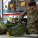 A U.S. Air Force airman named Trevor is working in what appears to be a supply or logistics warehouse, handling military duffel bags and packaged equipment on a work table. The airman is wearing OCP (Operational Camouflage Pattern) uniform with visible name tape 'TREVOR' and 'U.S. AIR FORCE' tape. The background shows industrial shelving with yellow tags, indicating an organized inventory or supply depot environment.