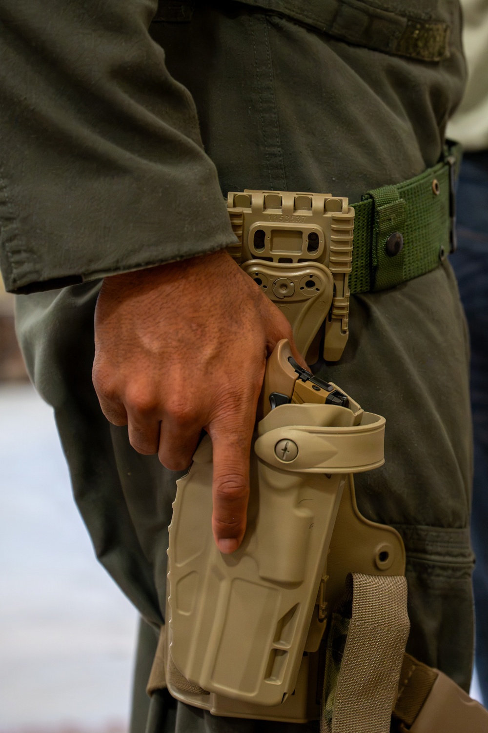A close-up image showing a person wearing olive/dark green military or tactical clothing with a tan/coyote-colored drop-leg holster system attached to a green nylon belt. The individual's right hand is resting on or near the holster, which contains what appears to be a semi-automatic pistol. The holster system features a modular mounting platform consistent with tactical duty holster designs.