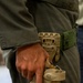 A close-up image showing a person wearing olive/dark green military or tactical clothing with a tan/coyote-colored drop-leg holster system attached to a green nylon belt. The individual's right hand is resting on or near the holster, which contains what appears to be a semi-automatic pistol. The holster system features a modular mounting platform consistent with tactical duty holster designs.