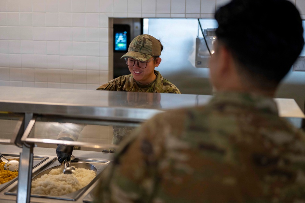 A military service member in camouflage uniform and cap is serving food at a cafeteria-style serving line, smiling while scooping what appears to be rice. Another service member in the foreground, seen from behind, is being served. The setting appears to be a military dining facility (DFAC).
