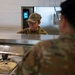 A military service member in camouflage uniform and cap is serving food at a cafeteria-style serving line, smiling while scooping what appears to be rice. Another service member in the foreground, seen from behind, is being served. The setting appears to be a military dining facility (DFAC).