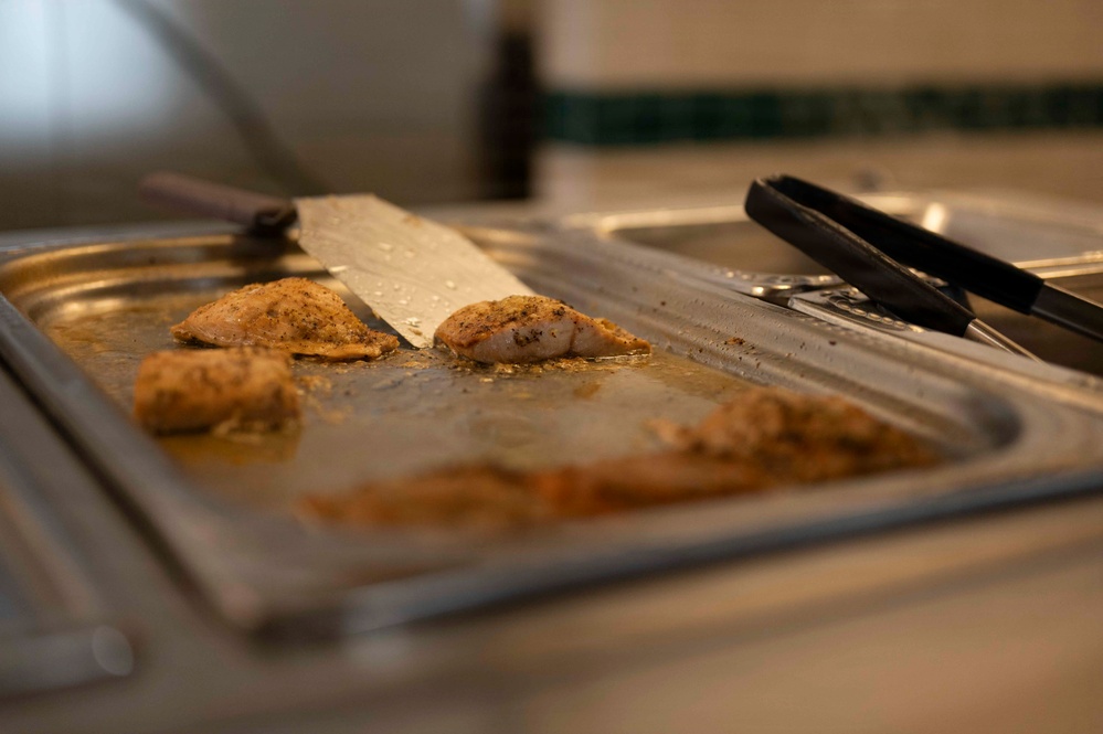 The image shows cooked seasoned chicken pieces on a stainless steel serving tray or hotel pan. A large chef's knife (cleaver-style) rests across the chicken, and black tongs are visible on the right side of the tray. The setting appears to be a commercial kitchen or food service line.