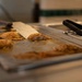 The image shows cooked seasoned chicken pieces on a stainless steel serving tray or hotel pan. A large chef's knife (cleaver-style) rests across the chicken, and black tongs are visible on the right side of the tray. The setting appears to be a commercial kitchen or food service line.