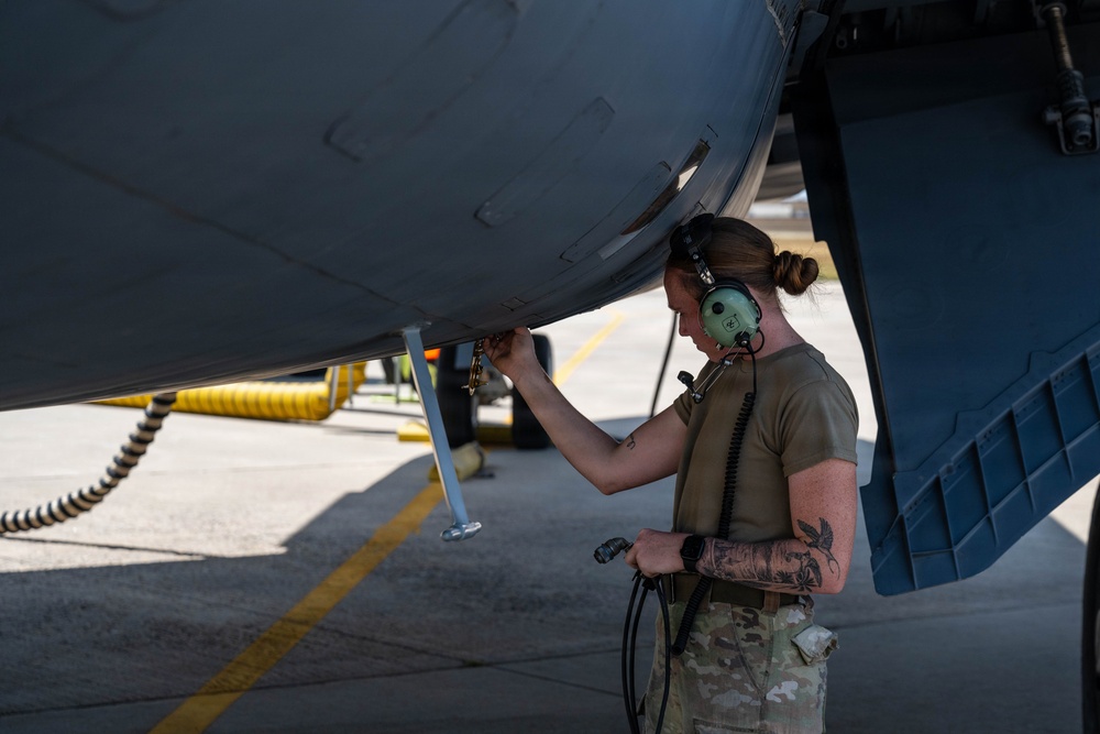 A female military aircraft maintainer in multicam/OCP uniform is performing maintenance work on the underside of a military aircraft on a flight line. She is wearing green aviation headset/hearing protection and has visible tattoos on her forearm. The setting appears to be an active airfield ramp area during daylight hours.