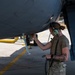 A female military aircraft maintainer in multicam/OCP uniform is performing maintenance work on the underside of a military aircraft on a flight line. She is wearing green aviation headset/hearing protection and has visible tattoos on her forearm. The setting appears to be an active airfield ramp area during daylight hours.