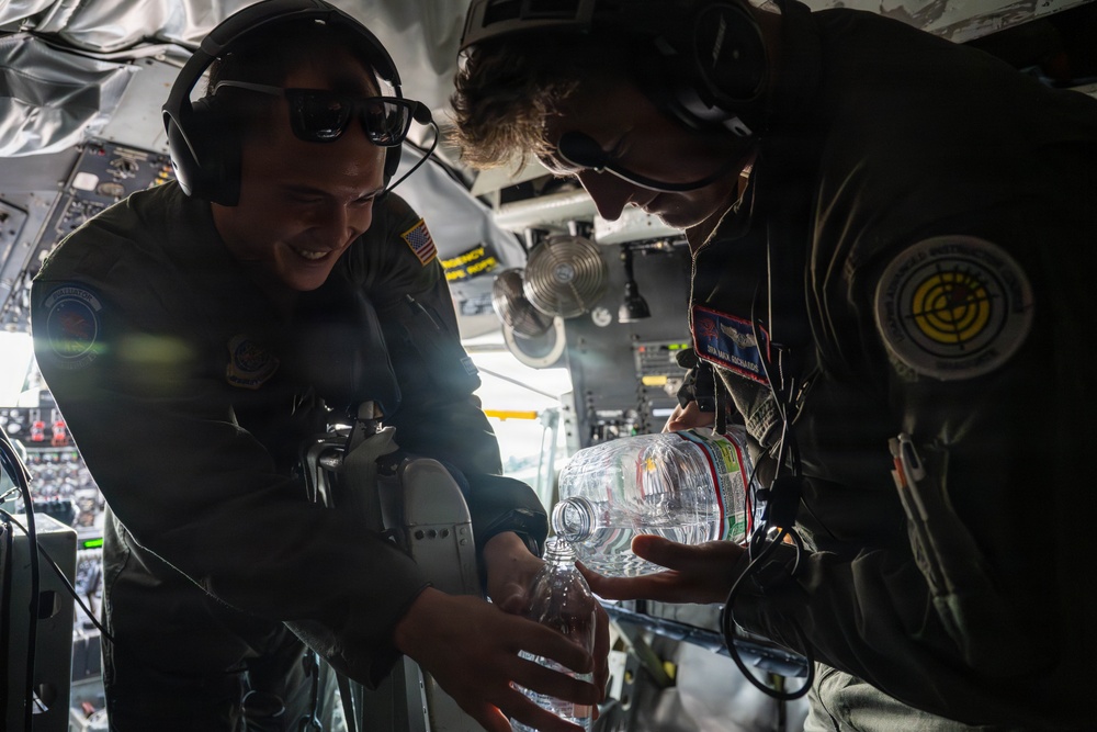 Two military aircrew members in flight suits are inside what appears to be a military aircraft, sharing or examining a large plastic water bottle or jug. Both individuals are wearing aviation headsets and patches on their flight suits, with one displaying a visible U.S. flag patch. The interior of the aircraft is visible in the background, including instrument panels and overhead equipment.