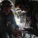 Two military aircrew members in flight suits are inside what appears to be a military aircraft, sharing or examining a large plastic water bottle or jug. Both individuals are wearing aviation headsets and patches on their flight suits, with one displaying a visible U.S. flag patch. The interior of the aircraft is visible in the background, including instrument panels and overhead equipment.