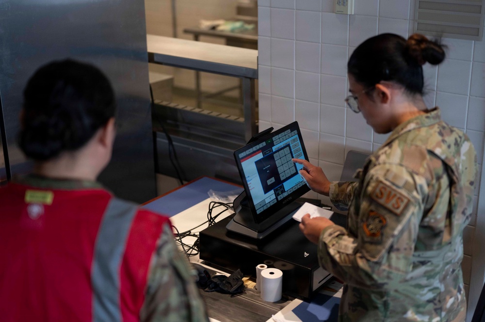 Two military personnel are working at what appears to be a food service point-of-sale station in a dining facility. One individual in camouflage uniform with 'SVS' patch is operating a touchscreen POS terminal, while another in a red vest observes. The setting appears to be a military dining facility or DFAC (Dining Facility).