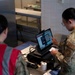 Two military personnel are working at what appears to be a food service point-of-sale station in a dining facility. One individual in camouflage uniform with 'SVS' patch is operating a touchscreen POS terminal, while another in a red vest observes. The setting appears to be a military dining facility or DFAC (Dining Facility).