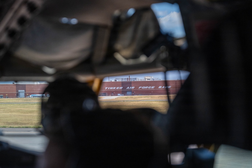 The image is taken from beneath or inside a large aircraft, looking through the undercarriage toward a large red brick building labeled 'Tinker Air Force Base.' The perspective is framed by mechanical components of the aircraft, with silhouetted figures visible in the foreground. The composition creates a tunnel-like view toward the base facility across an open tarmac area.