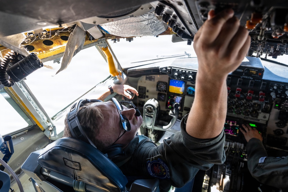 Two aircrew members are visible inside a military aircraft cockpit, with one pilot wearing a flight suit, headset, and aviator sunglasses looking upward toward the overhead panel. The cockpit displays a mix of analog gauges and some modern digital instruments, consistent with a legacy military aircraft such as a KC-135 or similar tanker/transport. The overhead panel shows signs of wear and aging, with visible corrosion and peeling insulation material.