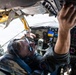 Two aircrew members are visible inside a military aircraft cockpit, with one pilot wearing a flight suit, headset, and aviator sunglasses looking upward toward the overhead panel. The cockpit displays a mix of analog gauges and some modern digital instruments, consistent with a legacy military aircraft such as a KC-135 or similar tanker/transport. The overhead panel shows signs of wear and aging, with visible corrosion and peeling insulation material.