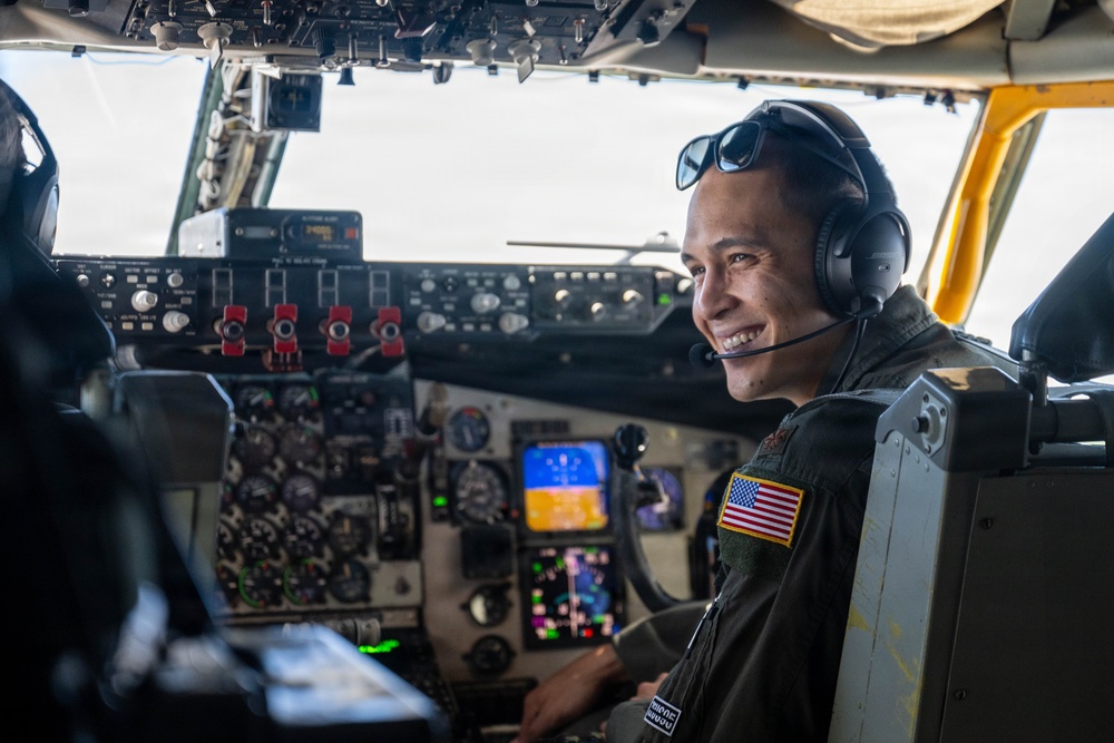 A smiling military aviator is seated in the cockpit of a large aircraft, wearing a flight suit with a U.S. flag patch, aviation headset, and sunglasses pushed up on his head. The cockpit features an extensive array of analog and digital instruments, switches, and displays consistent with a large military transport or tanker aircraft. The image is a portrait-style photograph taken from inside the cockpit.