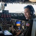 A smiling military aviator is seated in the cockpit of a large aircraft, wearing a flight suit with a U.S. flag patch, aviation headset, and sunglasses pushed up on his head. The cockpit features an extensive array of analog and digital instruments, switches, and displays consistent with a large military transport or tanker aircraft. The image is a portrait-style photograph taken from inside the cockpit.