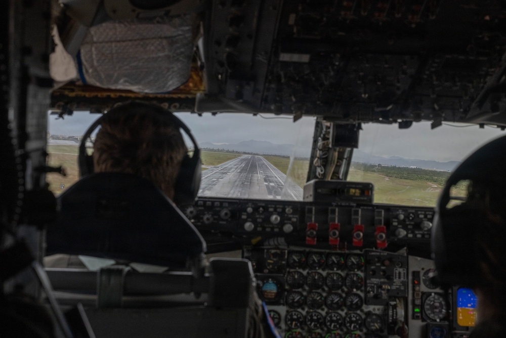 The image shows the interior of an aircraft cockpit from behind, with a pilot seated at the controls during what appears to be a takeoff or landing roll on a wet runway. The cockpit features an older-generation analog instrument panel with numerous gauges, switches, and red-guarded controls. Mountains and green terrain are visible in the background through the windscreen.
